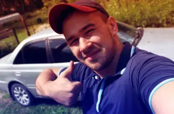 Man with hat, thumb raised, and grey car behind him 