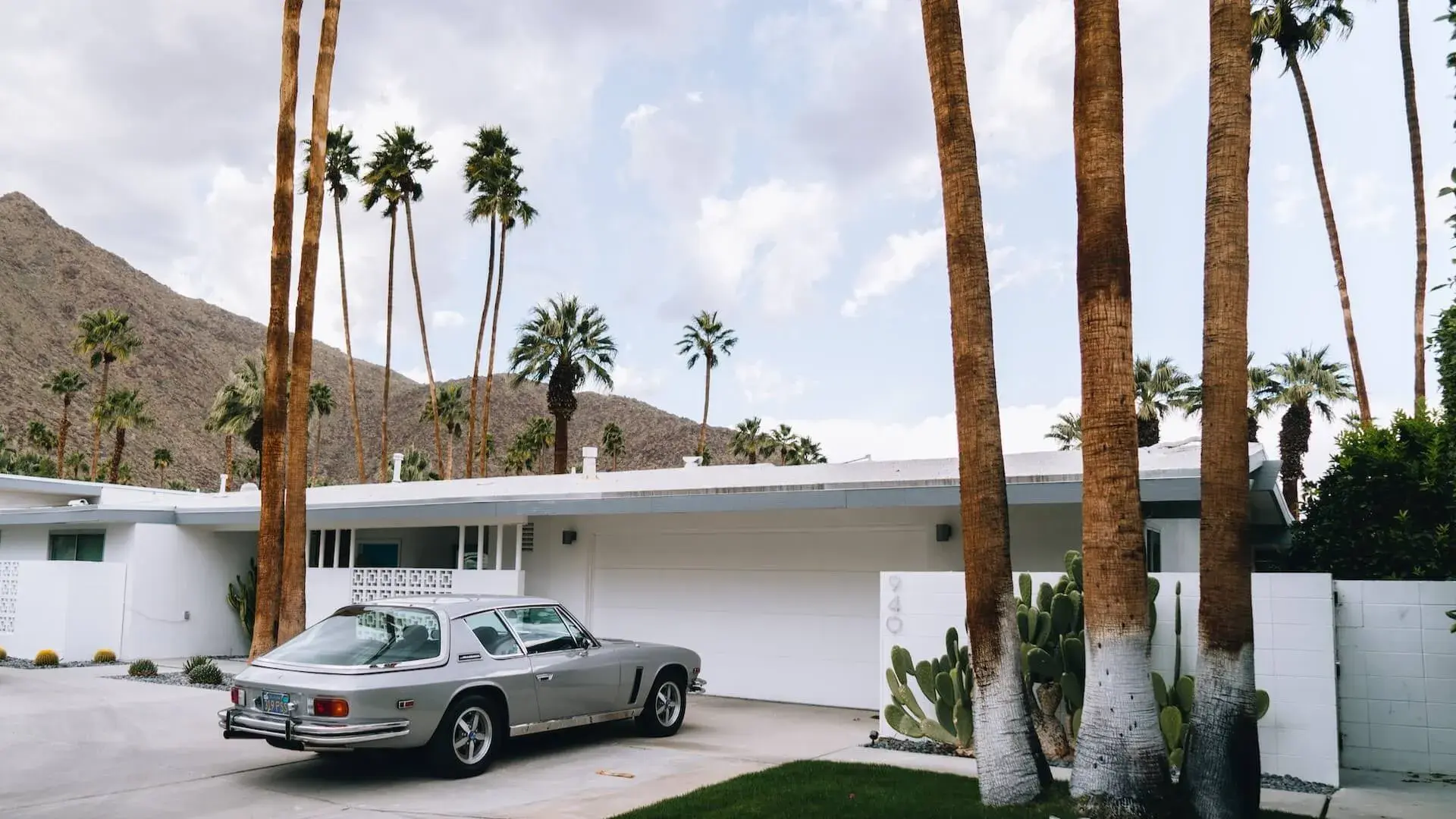 A grey car parked in front of a white big house with palms on the background
